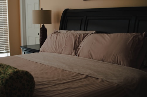A neatly made bed with light brown sheets and pillows, situated next to a dark-colored side table with a decorative lamp. The room features soft lighting and neutral wall colors. A door and window with blinds are partially visible.
