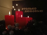 Four red candles are lit and placed on a decorative surface with green foliage and white berries. The background is dimly lit with hanging bulbs and white text in Russian.
