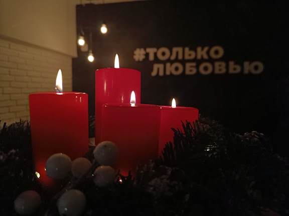 Four red candles are lit and placed on a decorative surface with green foliage and white berries. The background is dimly lit with hanging bulbs and white text in Russian.