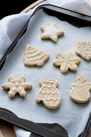 A baking tray with decorated sugar cookies in various festive shapes including a star, snowflake, Christmas tree, mitten, and snowman, placed on parchment paper. The cookies are outlined with white icing, providing a simple yet classic holiday appearance.