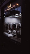 A dark exterior view of a dry cleaning and tailoring shop with large glass windows. Inside, racks of clothes covered in plastic are visible along with some bags and a chair. The lighting is dim, casting reflections on the glass.