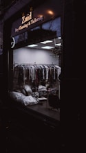 A dark exterior view of a dry cleaning and tailoring shop with large glass windows. Inside, racks of clothes covered in plastic are visible along with some bags and a chair. The lighting is dim, casting reflections on the glass.