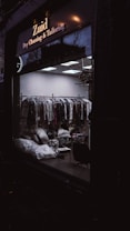 A dark exterior view of a dry cleaning and tailoring shop with large glass windows. Inside, racks of clothes covered in plastic are visible along with some bags and a chair. The lighting is dim, casting reflections on the glass.