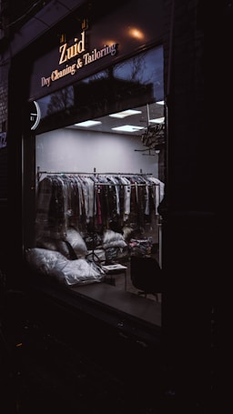 A dark exterior view of a dry cleaning and tailoring shop with large glass windows. Inside, racks of clothes covered in plastic are visible along with some bags and a chair. The lighting is dim, casting reflections on the glass.