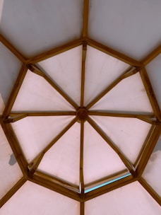 A wooden framed structure forms a geometric pattern resembling a dome. The beams create interconnected triangular sections, with a central polygon, allowing light to diffuse through the opaque panels.