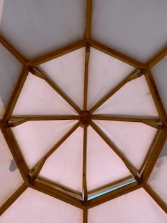 A wooden framed structure forms a geometric pattern resembling a dome. The beams create interconnected triangular sections, with a central polygon, allowing light to diffuse through the opaque panels.