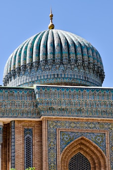 A highly ornate and intricately designed architectural dome, decorated with blue and turquoise tiles featuring detailed geometric and floral patterns. The structure is topped with a golden ornamental finial. The supporting walls include brickwork and further decorative tile work, combining traditional patterns and vibrant colors.