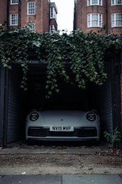 A front view of a white car parked inside a dimly lit garage. The car has a visible license plate reading 'NV19 MVF'. Above the garage entrance, green ivy plants hang down. Brick buildings with white-framed windows are visible in the background. The floor outside the garage is worn and has some scattered debris.