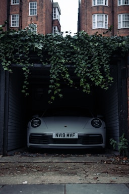 A front view of a white car parked inside a dimly lit garage. The car has a visible license plate reading 'NV19 MVF'. Above the garage entrance, green ivy plants hang down. Brick buildings with white-framed windows are visible in the background. The floor outside the garage is worn and has some scattered debris.