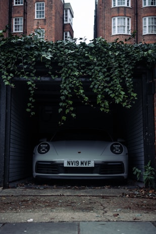 A front view of a white car parked inside a dimly lit garage. The car has a visible license plate reading 'NV19 MVF'. Above the garage entrance, green ivy plants hang down. Brick buildings with white-framed windows are visible in the background. The floor outside the garage is worn and has some scattered debris.