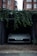 A front view of a white car parked inside a dimly lit garage. The car has a visible license plate reading 'NV19 MVF'. Above the garage entrance, green ivy plants hang down. Brick buildings with white-framed windows are visible in the background. The floor outside the garage is worn and has some scattered debris.