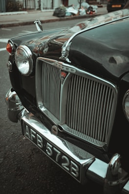 A vintage car with a chrome grille and distinctive emblem is parked on a city street. The license plate reads 'AFB 572B'. The headlights are round, and chrome elements accentuate the classic design. In the background, there are blurred hints of a sidewalk and some bags or debris.