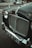 A vintage car with a chrome grille and distinctive emblem is parked on a city street. The license plate reads 'AFB 572B'. The headlights are round, and chrome elements accentuate the classic design. In the background, there are blurred hints of a sidewalk and some bags or debris.