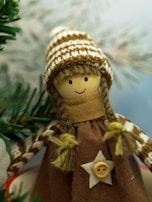 A handcrafted doll with a smiling face, wearing a knitted hat with brown and white stripes. It has braided hair made of yarn and wears a brown dress adorned with a button in the shape of a star. The background features blurred green pine branches, suggesting a festive or winter theme.