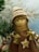 A handcrafted doll with a smiling face, wearing a knitted hat with brown and white stripes. It has braided hair made of yarn and wears a brown dress adorned with a button in the shape of a star. The background features blurred green pine branches, suggesting a festive or winter theme.