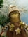 A handcrafted doll with a smiling face, wearing a knitted hat with brown and white stripes. It has braided hair made of yarn and wears a brown dress adorned with a button in the shape of a star. The background features blurred green pine branches, suggesting a festive or winter theme.