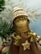 A handcrafted doll with a smiling face, wearing a knitted hat with brown and white stripes. It has braided hair made of yarn and wears a brown dress adorned with a button in the shape of a star. The background features blurred green pine branches, suggesting a festive or winter theme.