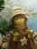 A handcrafted doll with a smiling face, wearing a knitted hat with brown and white stripes. It has braided hair made of yarn and wears a brown dress adorned with a button in the shape of a star. The background features blurred green pine branches, suggesting a festive or winter theme.