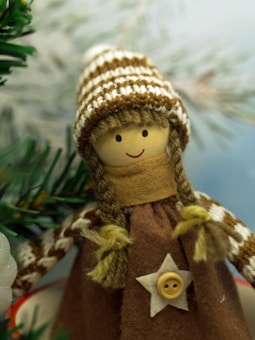 A handcrafted doll with a smiling face, wearing a knitted hat with brown and white stripes. It has braided hair made of yarn and wears a brown dress adorned with a button in the shape of a star. The background features blurred green pine branches, suggesting a festive or winter theme.