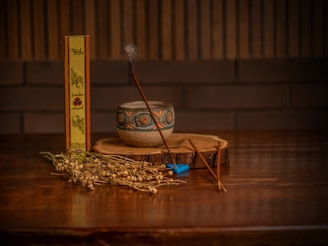 A wooden table holds a small, ornate ceramic bowl and a slender incense stick emitting a gentle wisp of smoke. Next to the bowl lies a dried plant arrangement and an incense box with decorative text and designs. A small, blue accessory rests near the incense stick, while a rustic wooden slab serves as a base.