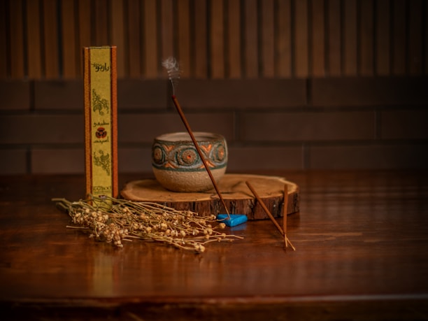 A wooden table holds a small, ornate ceramic bowl and a slender incense stick emitting a gentle wisp of smoke. Next to the bowl lies a dried plant arrangement and an incense box with decorative text and designs. A small, blue accessory rests near the incense stick, while a rustic wooden slab serves as a base.