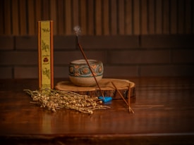A wooden table holds a small, ornate ceramic bowl and a slender incense stick emitting a gentle wisp of smoke. Next to the bowl lies a dried plant arrangement and an incense box with decorative text and designs. A small, blue accessory rests near the incense stick, while a rustic wooden slab serves as a base.