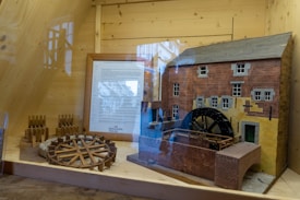 A detailed model of a historical watermill is displayed inside a wooden cabinet. The miniature depicts a brick building with multiple windows and a large black waterwheel attached to its side. There is a plaque with information beside the model, and various wooden components are arranged around it, possibly parts of the mill's mechanism.