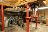 A mechanical setup featuring large interlocking gears supported by red metal pillars. The space has a rustic feel with wooden beams and a brick floor. The machinery appears to be part of an old mill or industrial setting, with visible wear indicating age and usage.