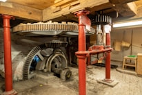 A mechanical setup featuring large interlocking gears supported by red metal pillars. The space has a rustic feel with wooden beams and a brick floor. The machinery appears to be part of an old mill or industrial setting, with visible wear indicating age and usage.