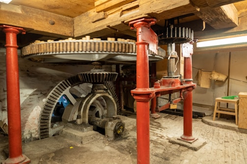 A mechanical setup featuring large interlocking gears supported by red metal pillars. The space has a rustic feel with wooden beams and a brick floor. The machinery appears to be part of an old mill or industrial setting, with visible wear indicating age and usage.