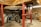A mechanical setup featuring large interlocking gears supported by red metal pillars. The space has a rustic feel with wooden beams and a brick floor. The machinery appears to be part of an old mill or industrial setting, with visible wear indicating age and usage.