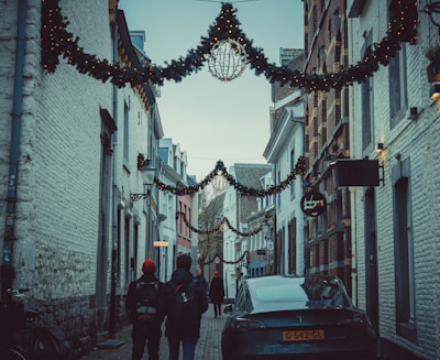 A narrow European street adorned with festive garlands and lights stretches between rows of old buildings. People wearing winter clothing walk along the cobblestone path, and a parked car is visible on the right side. The buildings are constructed from brick and stone, creating a cozy and historic atmosphere.