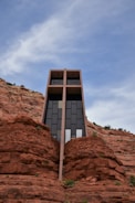 A modern architectural structure is built into a red rock cliff, featuring large glass windows and a striking geometric design. The building's vertical lines contrast with the natural, rugged texture of the surrounding rock formations under a blue sky with light clouds.