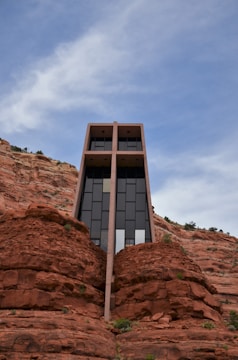 A modern architectural structure is built into a red rock cliff, featuring large glass windows and a striking geometric design. The building's vertical lines contrast with the natural, rugged texture of the surrounding rock formations under a blue sky with light clouds.