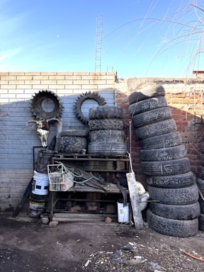 A collection of worn and used tires is stacked unevenly against a brick wall. There are also rims, a skull mounted on the wall, a tangle of hoses, and a metal rack with various tools and containers. The environment appears dusty and industrial, suggesting a storage or scrapyard setting.