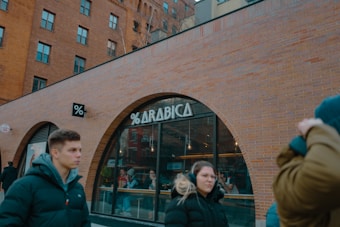A street scene featuring a brick building with large arched windows, one displaying the sign '% ARABICA'. Several people in warm clothing walk past, some looking towards the camera. Inside the cafe, patrons can be seen sitting at tables.