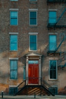 A brick facade of a multi-story building featuring several windows and a prominent red door with the address '8 Old Fulton St'. The entryway is accessed by a set of black metal stairs. Fire escape ladders are visible on the side of the building.