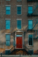 A brick facade of a multi-story building featuring several windows and a prominent red door with the address '8 Old Fulton St'. The entryway is accessed by a set of black metal stairs. Fire escape ladders are visible on the side of the building.