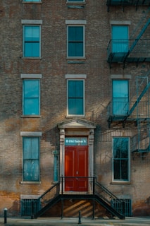 A brick facade of a multi-story building featuring several windows and a prominent red door with the address '8 Old Fulton St'. The entryway is accessed by a set of black metal stairs. Fire escape ladders are visible on the side of the building.