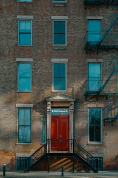 A brick facade of a multi-story building featuring several windows and a prominent red door with the address '8 Old Fulton St'. The entryway is accessed by a set of black metal stairs. Fire escape ladders are visible on the side of the building.