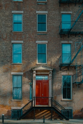 A brick facade of a multi-story building featuring several windows and a prominent red door with the address '8 Old Fulton St'. The entryway is accessed by a set of black metal stairs. Fire escape ladders are visible on the side of the building.