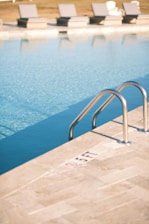 A calm swimming pool with clear blue water, featuring a metal pool ladder on the right side. The poolside area is tiled with light-colored stone, marked with a '5 FT' depth indicator. In the background, multiple cushioned sun loungers are aligned, with grassy elements visible beyond them.