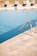 A calm swimming pool with clear blue water, featuring a metal pool ladder on the right side. The poolside area is tiled with light-colored stone, marked with a '5 FT' depth indicator. In the background, multiple cushioned sun loungers are aligned, with grassy elements visible beyond them.
