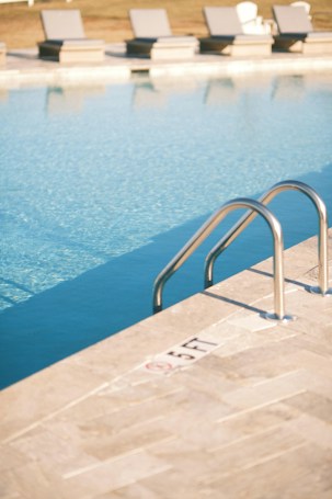 A calm swimming pool with clear blue water, featuring a metal pool ladder on the right side. The poolside area is tiled with light-colored stone, marked with a '5 FT' depth indicator. In the background, multiple cushioned sun loungers are aligned, with grassy elements visible beyond them.