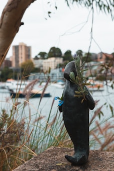 A small black sculpture resembling a standing animal is adorned with blue and purple flowers. The sculpture is placed on a stone surface with tall grasses around it. In the background, there is a blurry view of water, boats, and distant buildings, giving the scene an urban riverside feel.