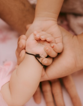 A delicate and tender scene where an adult hand gently holds a baby's hand, showcasing a sense of connection and care. The baby's wrist is adorned with a simple black string bracelet.