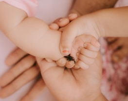 A close-up view of three hands, one belonging to a baby being gently held by an older child and an adult. The focus is on the tenderness and connection between the hands, with the baby's hand grasping a small black object.