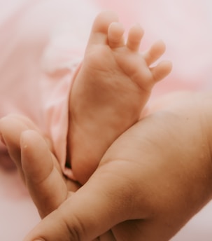 A close-up view of a baby's foot resting in an adult's hand, with soft lighting and a pink background, conveying a sense of tenderness and care.