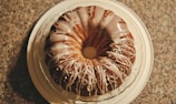A round bundt cake with a drizzle of white icing on top, placed on a white plate. The cake is set on a textured countertop, creating a contrast with its smooth icing. The icing is unevenly spread, adding a sense of homemade charm.