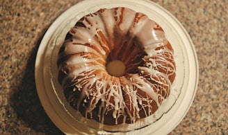 A round bundt cake with a drizzle of white icing on top, placed on a white plate. The cake is set on a textured countertop, creating a contrast with its smooth icing. The icing is unevenly spread, adding a sense of homemade charm.